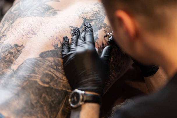 un hombre profesional con una máquina de tatuaje pone un tatuaje en la espalda de un hombre, de cerca. Tatuaje artista en el flujo de trabajo - Foto, Imagen