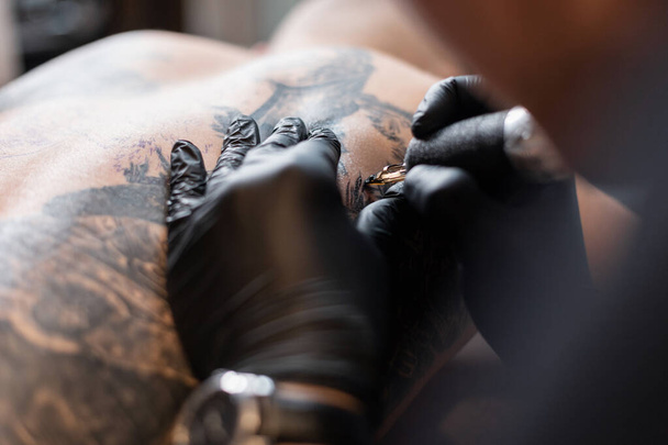 Tatuaje artista profesional hombre con guantes negros y una máquina de tatuaje dibuja con tinta en el cuerpo, primer plano. El proceso de trabajo - Foto, Imagen