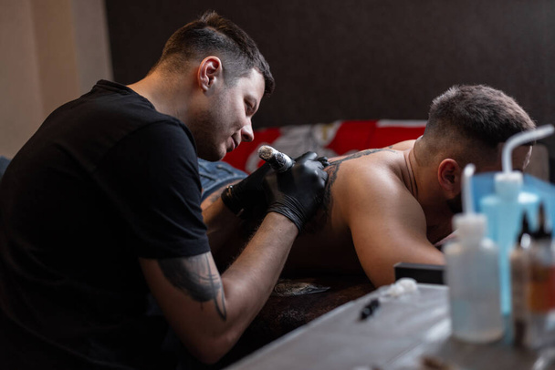 Hipster homem tatuagem mestre profissional com uma máquina de tatuagem faz um desenho nas costas de um homem no estúdio. Fluxo de trabalho de recheio de tatuagem - Foto, Imagem