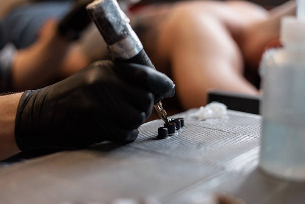 Profi férfi tetováló művész egy fekete kesztyű egy tetováló gép felveszi tinta, és egy tetoválás, közelkép - Fotó, kép