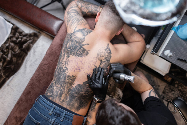 Profi tetoválóművész férfi fekete kesztyűvel és tetoválógép tetoválással egy férfi hátán, felülnézetből. Rajzolási munkafolyamat - Fotó, kép