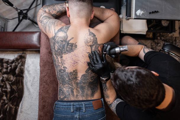 Homem artista de tatuagem com luvas pretas desenha uma tatuagem de cavaleiro nas costas do cara no estúdio, vista superior. Fluxo de trabalho do tatuador - Foto, Imagem