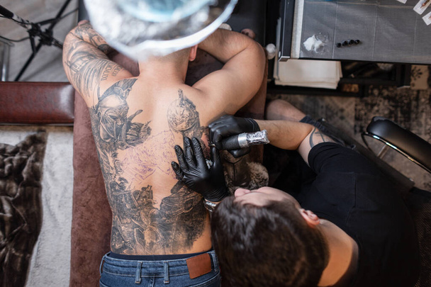 Man professional tattoo artist with black gloves and a modern tattoo machine makes a tattoo with ink on a male back, top view. tattoo creation workflow - Photo, Image