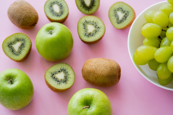 top view of grapes in bowl near kiwi and apples on pink  - Foto, Bild