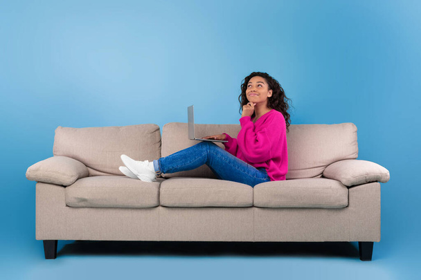 Στοχαστικό νεαρή μαύρη γυναίκα σε casual φορούν εργασίας σε απευθείας σύνδεση, κάθεται στον καναπέ και τη χρήση φορητού υπολογιστή σε μπλε φόντο στούντιο - Φωτογραφία, εικόνα