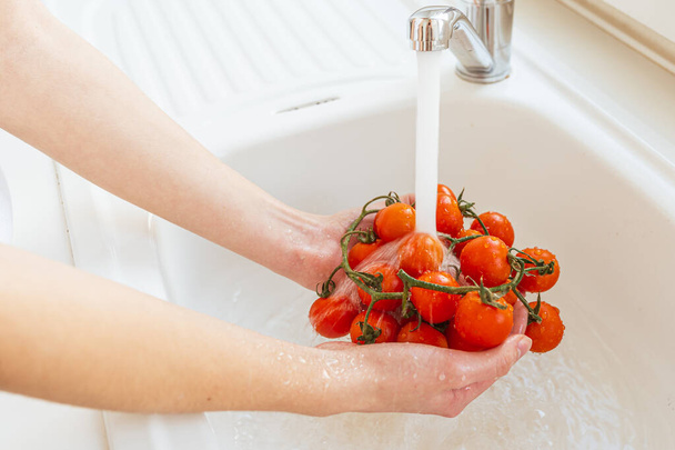 Fresh cherry tomatoes on bunch, rinse under running water in kitchen sink. Woman's hands holding freshly picked tomatoes under water. Drops of jet of water flow down from ripe red tomatoes, female hands wash fresh vegetables - Φωτογραφία, εικόνα