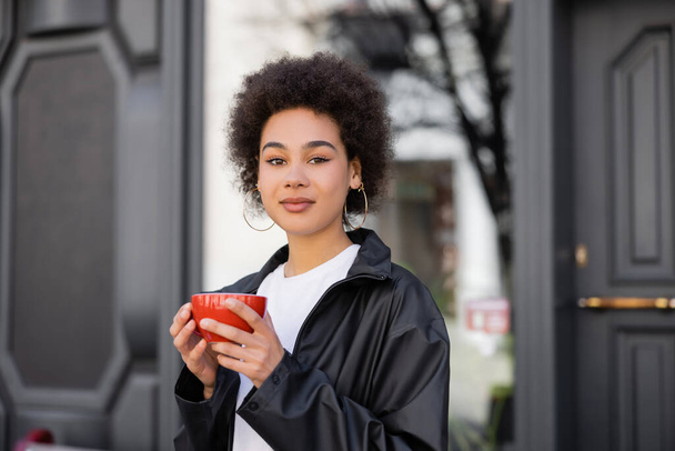 curly african american woman holding cup of coffee outside  - Foto, afbeelding