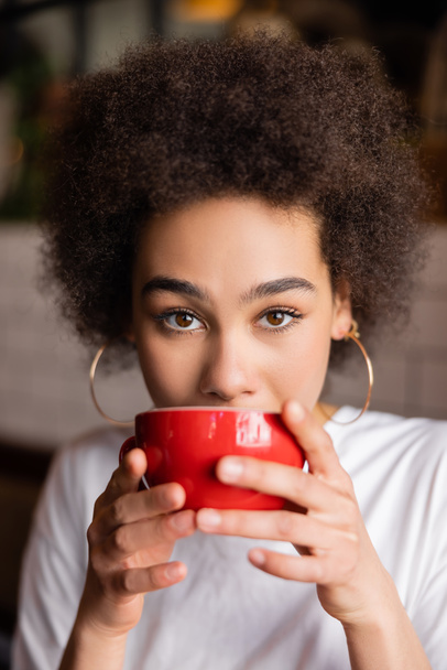 african american woman in hoop earrings drinking coffee and looking at camera  - Foto, imagen