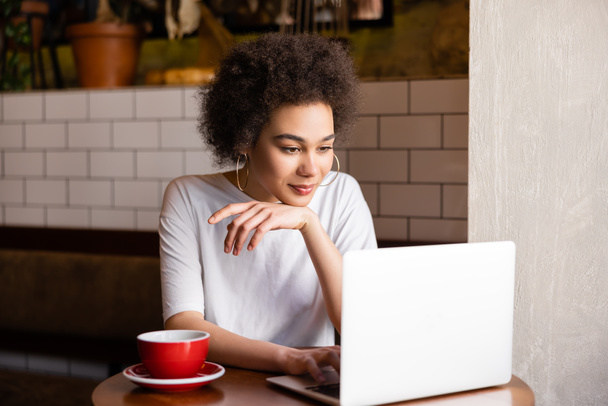 curly african american woman using laptop near cup of coffee in cafe  - Foto, immagini