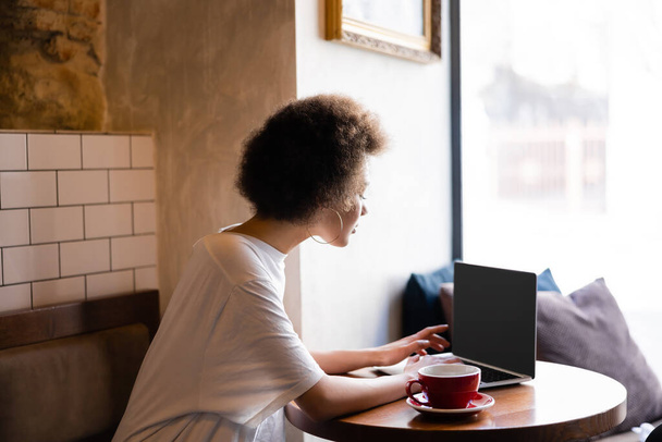 curly african american woman using laptop with blank screen near cup of coffee in cafe  - Foto, Imagen