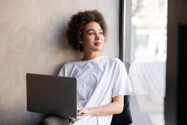 Beyaz tişörtlü genç Afro-Amerikalı kadın elinde laptop, pencerenin yanında.  - Fotoğraf, Görsel