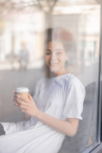 happy african american woman in white t-shirt holding paper cup behind window  - Fotografie, Obrázek