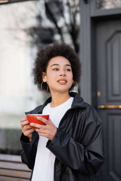 donna afro-americana in giacca con in mano una tazza di caffè fuori  - Foto, immagini