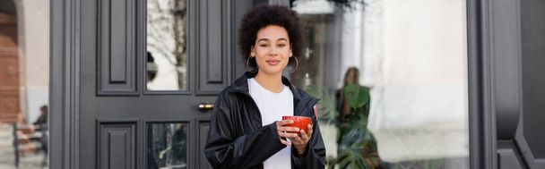 pleased african american woman in jacket holding cup of coffee on street, banner - Foto, Bild
