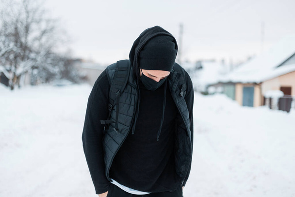 Fashionable handsome young hipster man in fashion black sports outerwear with hoodie, vest, hat and backpack walks on a winter day with snow in the countryside - 写真・画像