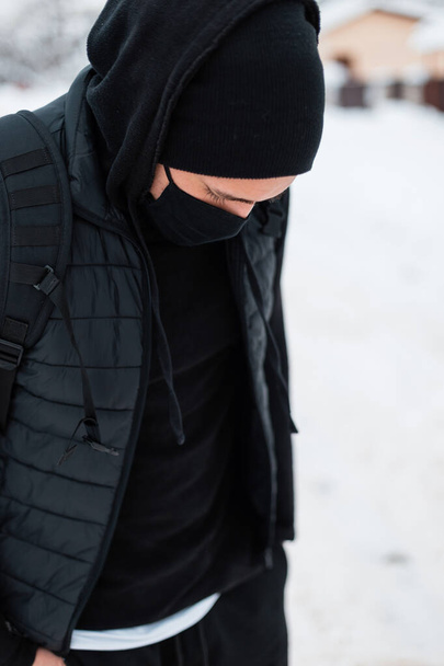 黒ベスト、パーカー、帽子とバックパックが冬の雪の日に歩くとファッショナブルな服で黒保護医療マスクを持つスタイリッシュなハンサムな若い男 - 写真・画像