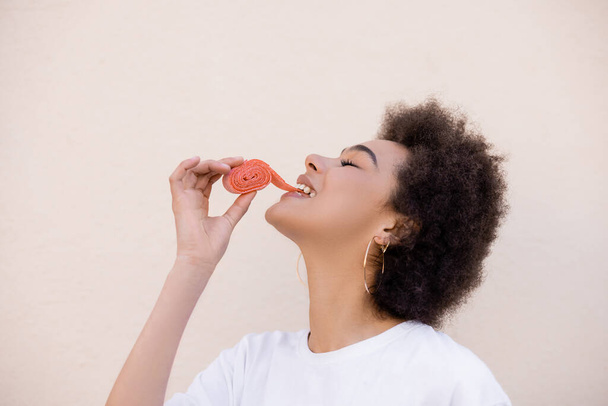happy african american young woman eating jelly bubble tape on white - Foto, afbeelding