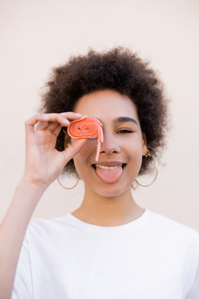 happy african american young woman covering eye with jelly bubble tape and sticking out tongue on white - Foto, imagen