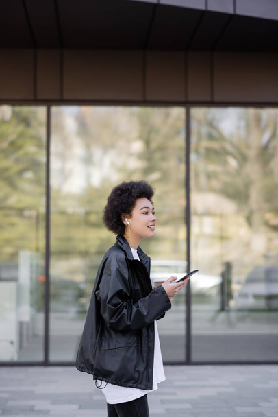 femme afro-américaine gaie en veste et écouteur tenant smartphone à l'extérieur  - Photo, image