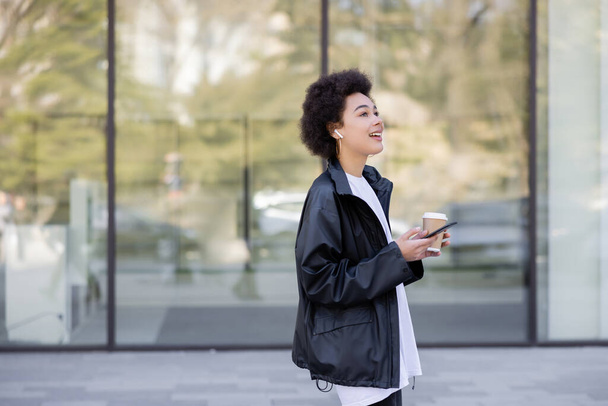 usmívající se africká americká žena ve sluchátku drží odnést nápoj a smartphone venku - Fotografie, Obrázek