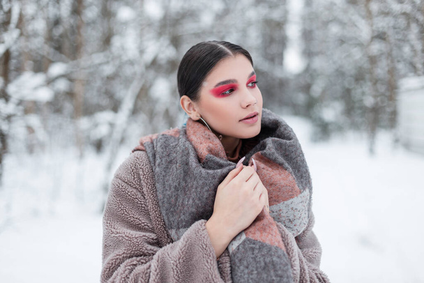 beautiful young model woman with colored bright makeup and pink eye shadows in fashionable winter clothes with a fur coat and scarf has against the background of a snow forest - Photo, Image