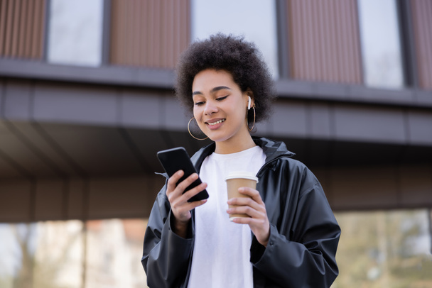 low angle view of smiling african american woman in earphone holding paper cup and smartphone outside - Zdjęcie, obraz