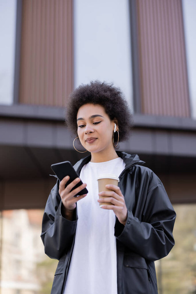 low angle view of curly african american woman in earphone holding paper cup and smartphone outside - Fotó, kép