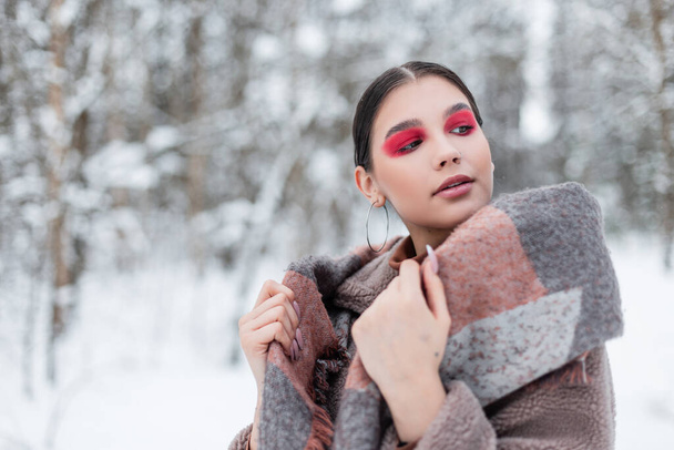 Mujer creativa joven y elegante con sombras de ojos rosados en ropa de lana de moda con una bufanda vintage en un frío día de invierno con nieve - Foto, Imagen