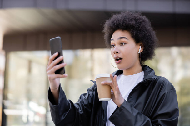 šokovaný africký Američan žena ve sluchátku drží papírový kelímek a mají video chat na smartphone venku - Fotografie, Obrázek