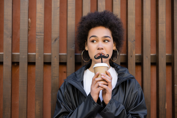 curly african american woman drinking takeaway beverage from straw with moustache near fence - Valokuva, kuva