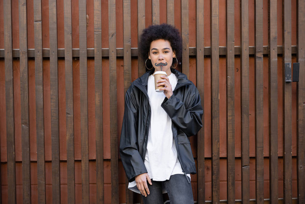 giovane donna afroamericana che beve bevanda da asporto dalla paglia con i baffi vicino alla recinzione - Foto, immagini