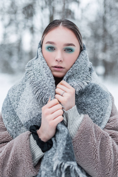 Χειμερινό πορτραίτο ενός όμορφου νεαρού Ρώσου μοντέλου με μοντέρνα χειμωνιάτικα ρούχα με ένα vintage ζεστό μαντήλι, γούνινο παλτό και ένα πουλόβερ στέκεται στο δάσος με χιόνι - Φωτογραφία, εικόνα