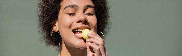 pleased african american woman eating yellow candy, banner - Zdjęcie, obraz