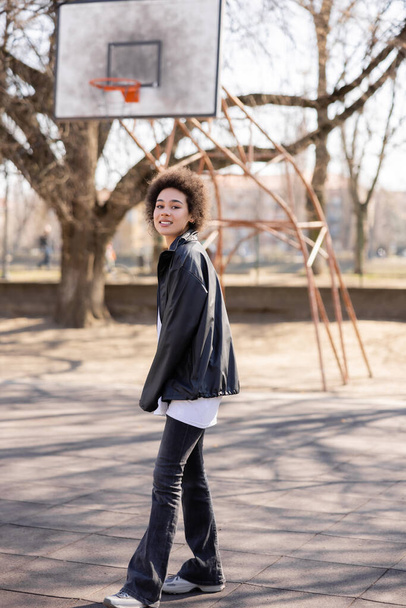 full length view of happy african american woman in jacket near basketball outdoor court - Foto, immagini