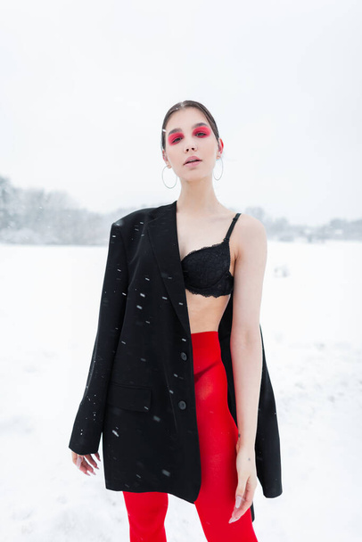 Όμορφο μοντέλο εσώρουχα κορίτσι σε μοντέρνα ρούχα με κόκκινο καλσόν, σακάκι και σουτιέν θέτουν σε εξωτερικούς χώρους με χιόνι στη φύση - Φωτογραφία, εικόνα