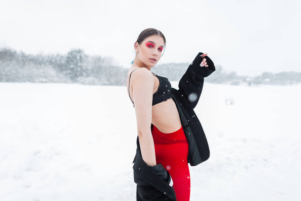 Λαμπερό σέξι νεαρή Vogue μοντέλο κορίτσι με φωτεινό μακιγιάζ σε vintage ρούχα με κόκκινο καλσόν μόδας, ένα σακάκι και ένα μαύρο σουτιέν ποζάρουν στη φύση με χιόνι - Φωτογραφία, εικόνα