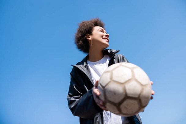 low angle view of cheerful african american woman in jacket holding ball against blue sky - Foto, Bild