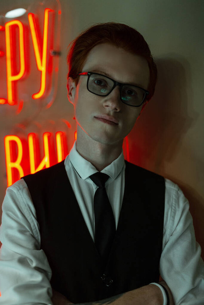 Retrato moderno de um jovem hipster bonito com óculos em roupas de negócios com uma camisa, colete e gravata fica perto de uma parede verde com novas luzes vermelhas - Foto, Imagem