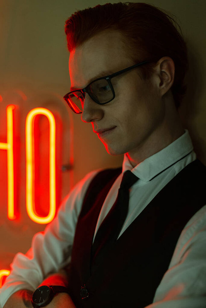 Μόδα έγχρωμο πορτρέτο ενός όμορφου νεαρού hipster άνδρα με γυαλιά και χτένισμα σε κομψά επαγγελματικά ρούχα με κοστούμι και γραβάτα στέκεται κοντά στον τοίχο με πράσινο και κόκκινο νέον φώτα. - Φωτογραφία, εικόνα