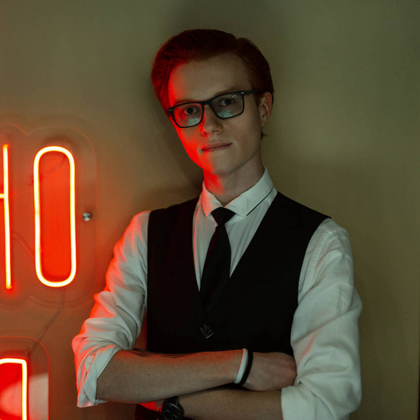 Negocio joven pelirrojo guapo con gafas en ropa de moda con un traje y corbata se encuentra cerca de una pared verde con luz de neón rojo - Foto, Imagen
