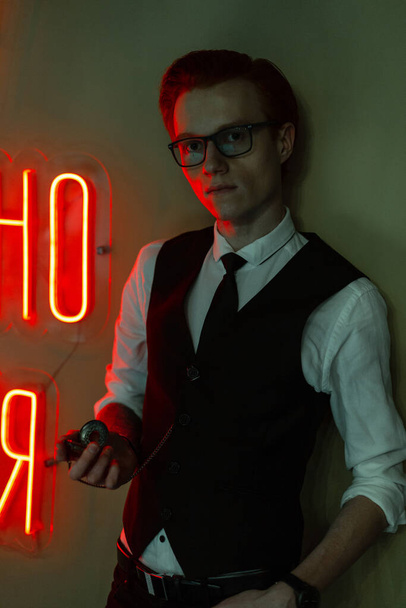 Κομψός όμορφος νεαρός επιχειρηματίας μοντέλο με γυαλιά σε μοντέρνα επίσημα ρούχα με πουκάμισο, γιλέκο και γραβάτα με ρολόι κοντά σε ένα πράσινο τοίχο με κόκκινο φως νέον. Χρόνος, εργασία και γραφείο - Φωτογραφία, εικόνα