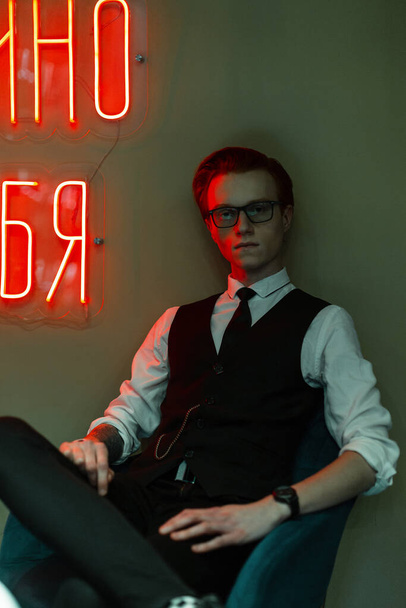 El inglés guapo a la moda pelirrojo con gafas en ropa de negocios con un traje y corbata se sienta en una silla cerca de la pared verde con letras rojas de neón - Foto, imagen