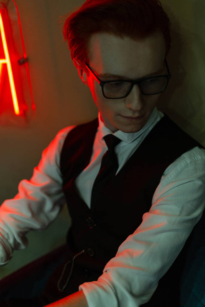 Homem ruivo bonito elegante com óculos em roupas de negócios de moda com uma camisa, colete e gravata fica perto de uma parede verde em luz vermelha. Estilo masculino de negócios - Foto, Imagem