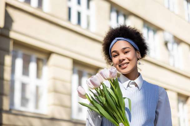 sonriente afroamericana mujer sosteniendo tulipanes en la calle  - Foto, Imagen
