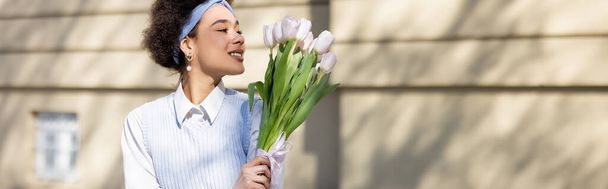 joyful african american woman looking at bouquet of tulips on street, banner - 写真・画像