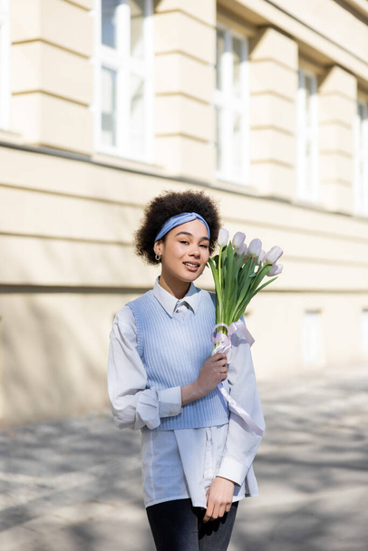 joven afroamericana mujer sosteniendo tulipanes y sonriendo en la calle  - Foto, imagen