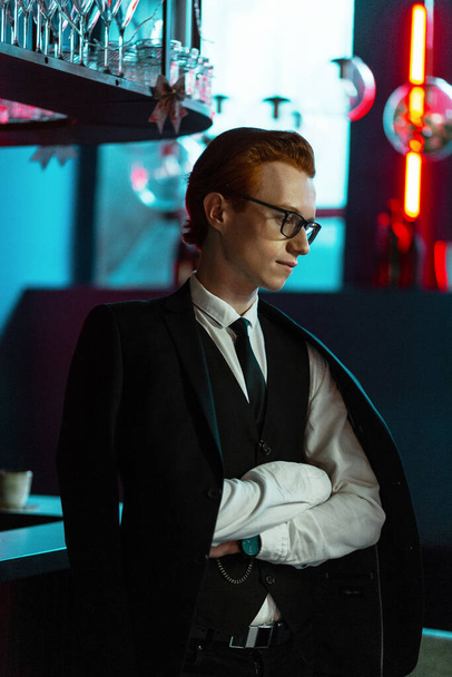 Joven hombre de negocios guapo de moda con gafas en ropa de negocios con estilo con un traje y corbata se encuentra en un bar con luz azul y roja - Foto, Imagen