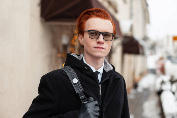 Elegante giovane uomo hipster rossa con gli occhiali in una giacca nera con uno zaino passeggiate in città. Viaggiatore d'affari in capispalla alla moda in una giornata invernale. Ritratto maschile - Foto, immagini