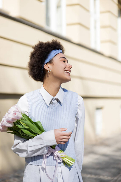 positive young african american woman in blue vest and white shirt holding bouquet of tulips - Foto, imagen