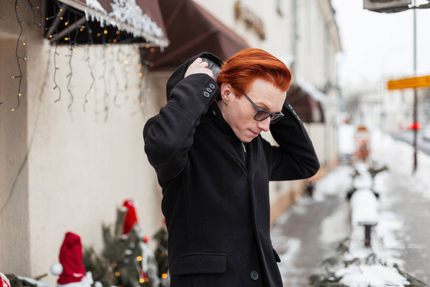 gutaussehender junger Geschäftsmann mit leuchtend roten Haaren in modischer Oberbekleidung mit Mantel trägt auf der Straße in der Stadt eine Kapuze. Englischer Männerstil im Freien - Foto, Bild
